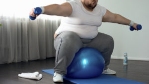 Corpulent male in sportswear lifting dumbbells sitting on fitness ball, activity